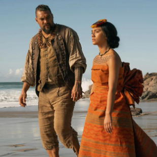 Actors Jason Mamoa and Luciane Buchanan wear period-style clothing and walk along a sandy beach near the ocean while filming Chief of War.