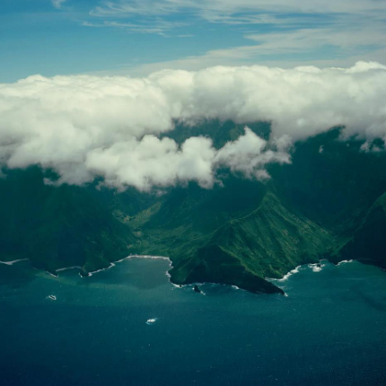 Aerial view of a lush, mountainous coastline with steep green cliffs descending into deep blue ocean waters, partially covered by low-hanging clouds.