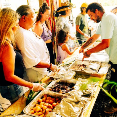 People stand in line at a buffet table outdoors as a server portions food onto plates. Trays of prepared dishes are arranged on the table, and several adults and a child wait to be served.