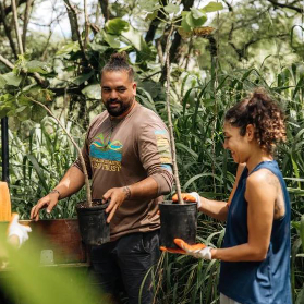Two people stand outdoors among tall plants, holding potted saplings and gardening tools, participating in a conservation activity.