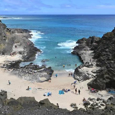 Secluded sandy cove surrounded by dark rocky cliffs, with people swimming in clear blue water and relaxing on the beach under a bright sky.