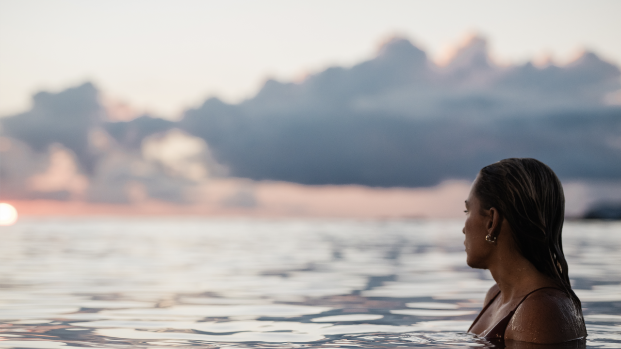 women chest deep in the ocean looking out over the horizon as the sun sets